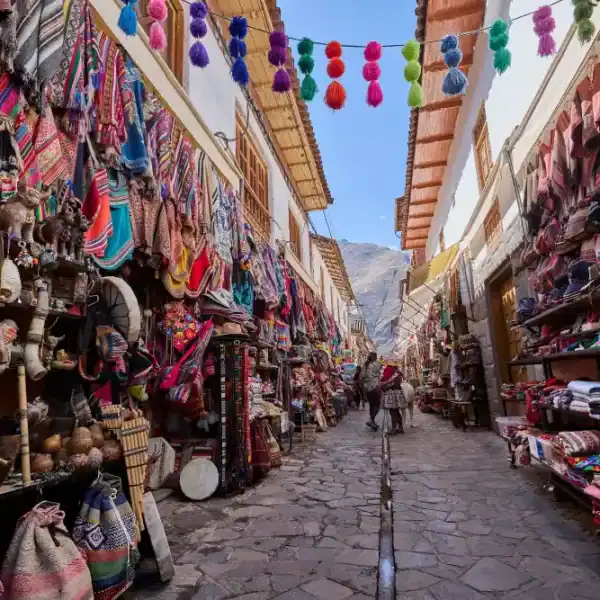 Rua estreita de pedra em um mercado tradicional andino, cercada por lojas repletas de artesanato colorido, tecidos, bolsas, chapéus e lembranças típicas. Pompons multicoloridos estão pendurados sobre a via, enquanto visitantes caminham pelo corredor com montanhas ao fundo sob céu azul.