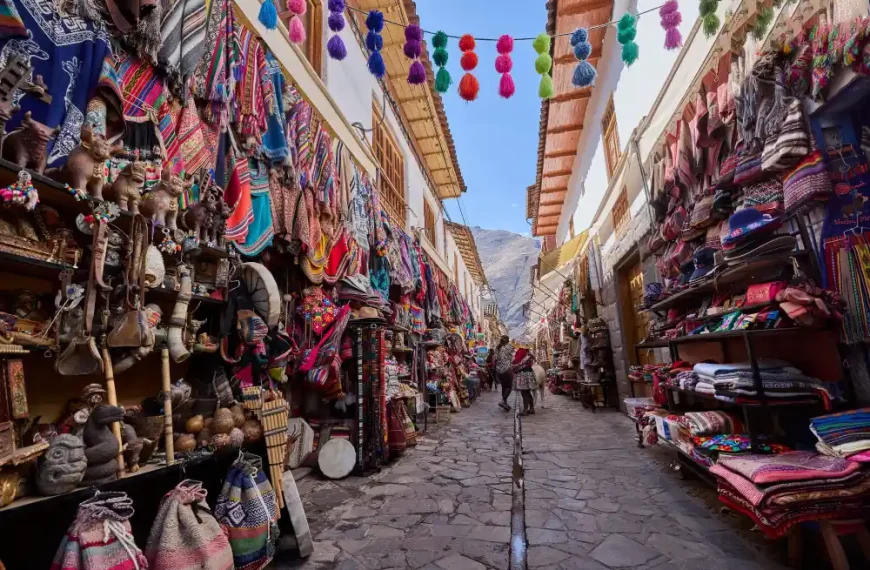 Rua estreita de pedra em um mercado tradicional andino, cercada por lojas repletas de artesanato colorido, tecidos, bolsas, chapéus e lembranças típicas. Pompons multicoloridos estão pendurados sobre a via, enquanto visitantes caminham pelo corredor com montanhas ao fundo sob céu azul.