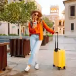 Mulher turista caminhando por uma praça de arquitetura histórica europeia ao entardecer, usando chapéu, óculos de sol, blusa laranja e calça jeans clara. Ela segura um smartphone enquanto apoia a mão em uma mala amarela de rodinhas. Ao redor, há árvores em grandes jardineiras de madeira e edifícios de fachada clara ao fundo, iluminados pela luz dourada do fim do dia.