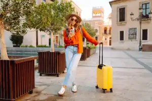 Mulher turista caminhando por uma praça de arquitetura histórica europeia ao entardecer, usando chapéu, óculos de sol, blusa laranja e calça jeans clara. Ela segura um smartphone enquanto apoia a mão em uma mala amarela de rodinhas. Ao redor, há árvores em grandes jardineiras de madeira e edifícios de fachada clara ao fundo, iluminados pela luz dourada do fim do dia.