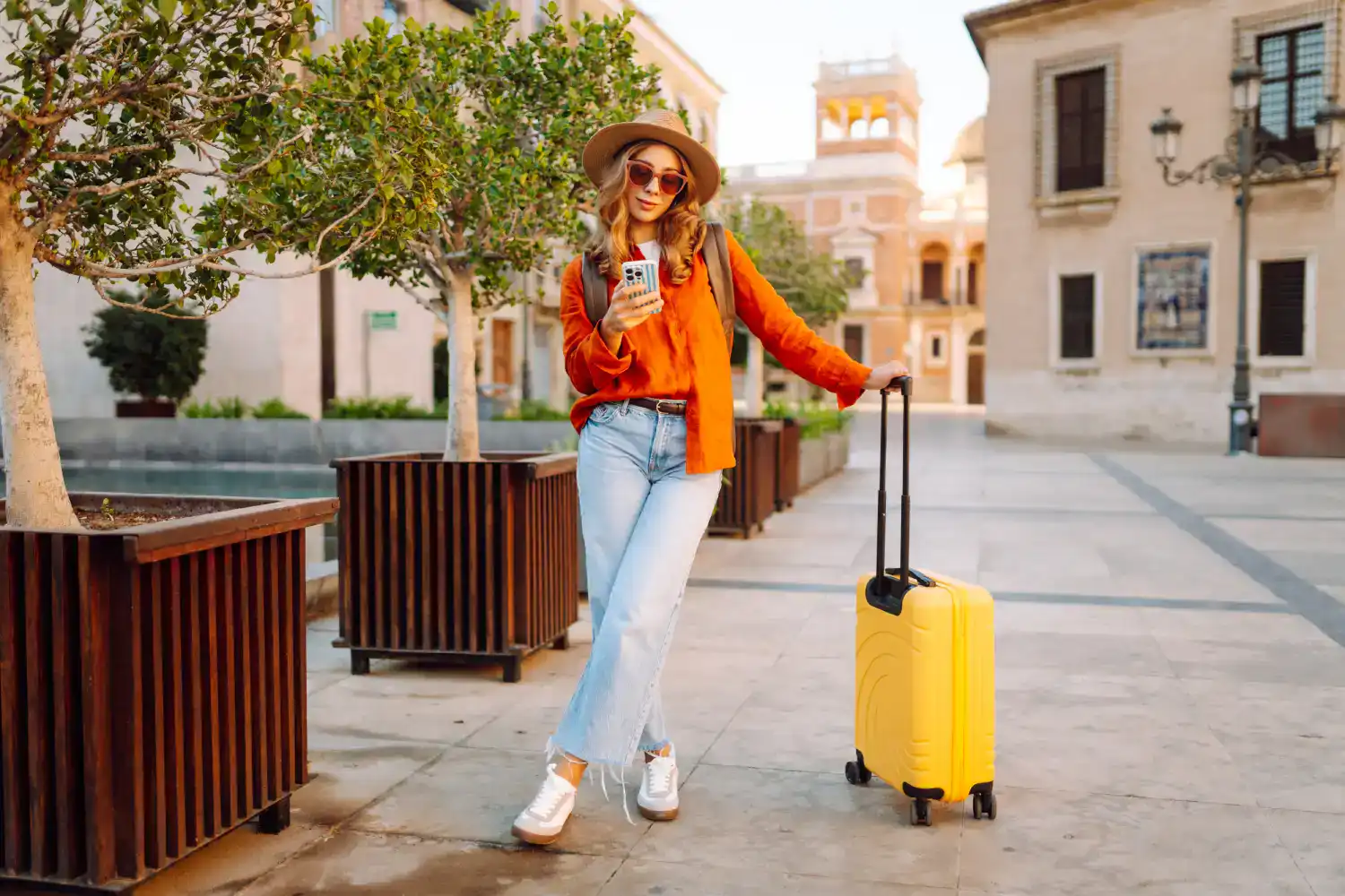 Mulher turista caminhando por uma praça de arquitetura histórica europeia ao entardecer, usando chapéu, óculos de sol, blusa laranja e calça jeans clara. Ela segura um smartphone enquanto apoia a mão em uma mala amarela de rodinhas. Ao redor, há árvores em grandes jardineiras de madeira e edifícios de fachada clara ao fundo, iluminados pela luz dourada do fim do dia.