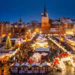 Vista panorâmica de um mercado de Natal iluminado em uma praça histórica coberta por neve, com barracas decoradas, árvore de Natal, carrossel e visitantes ao entardecer, cercados por edifícios históricos.