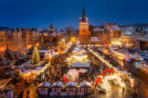 Vista panorâmica de um mercado de Natal iluminado em uma praça histórica coberta por neve, com barracas decoradas, árvore de Natal, carrossel e visitantes ao entardecer, cercados por edifícios históricos.
