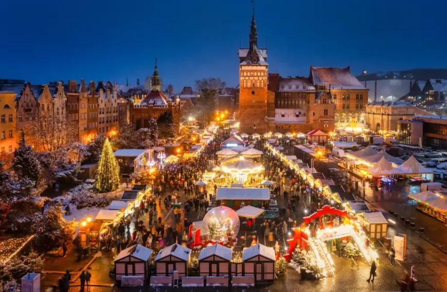Vista panorâmica de um mercado de Natal iluminado em uma praça histórica coberta por neve, com barracas decoradas, árvore de Natal, carrossel e visitantes ao entardecer, cercados por edifícios históricos.