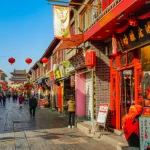 Rua comercial tradicional decorada com lanternas vermelhas chinesas, cercada por lojas, restaurantes e edifícios históricos, com pedestres caminhando em um bairro cultural durante o dia