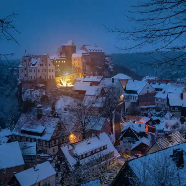 Uma pitoresca vila europeia de inverno coberta de neve ao anoitecer, destacando um castelo iluminado no topo de uma colina rochosa.