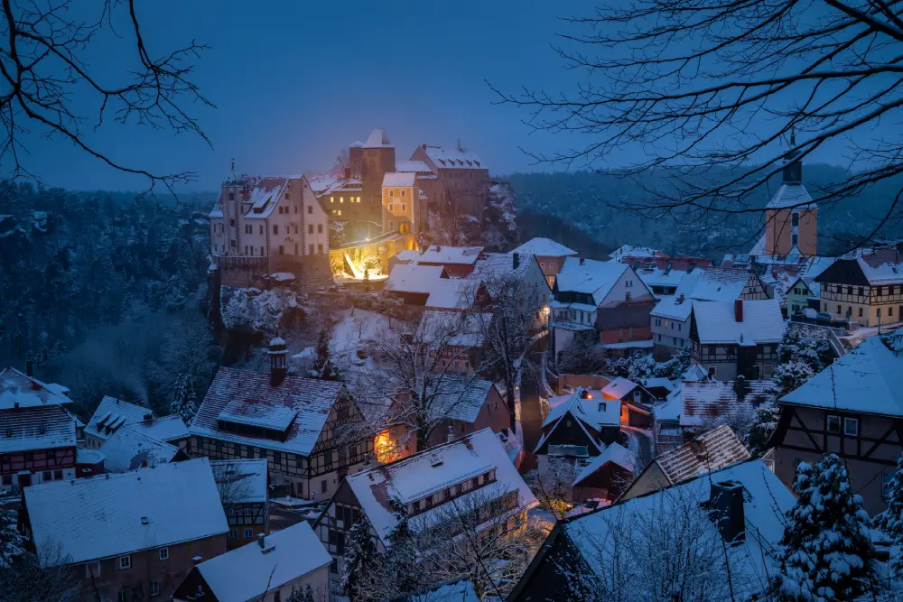 Uma pitoresca vila europeia de inverno coberta de neve ao anoitecer, destacando um castelo iluminado no topo de uma colina rochosa.