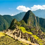 Vista panorâmica das antigas ruínas de pedra de Machu Picchu, com seus terraços agrícolas verdes espalhados pelo cume. Ao fundo, a icônica e íngreme montanha Huayna Picchu ergue-se majestosamente sob um céu azul com nuvens brancas, cercada por outros picos montanhosos da Cordilheira dos Andes.