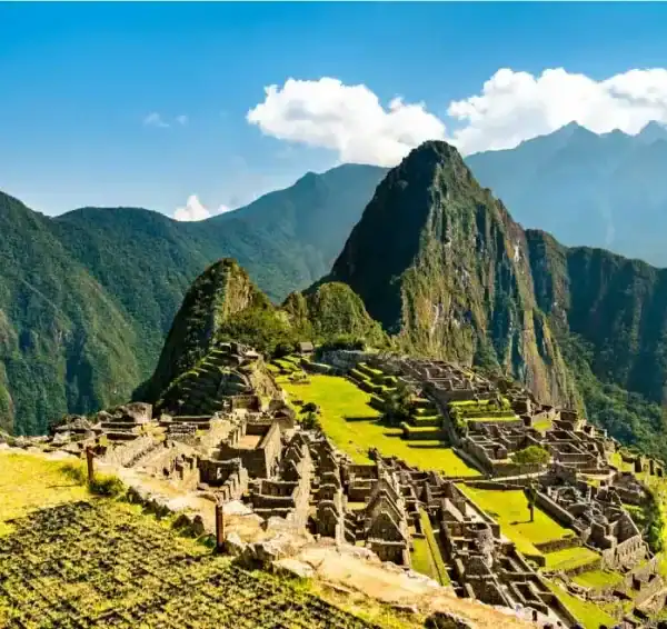 Vista panorâmica das antigas ruínas de pedra de Machu Picchu, com seus terraços agrícolas verdes espalhados pelo cume. Ao fundo, a icônica e íngreme montanha Huayna Picchu ergue-se majestosamente sob um céu azul com nuvens brancas, cercada por outros picos montanhosos da Cordilheira dos Andes.