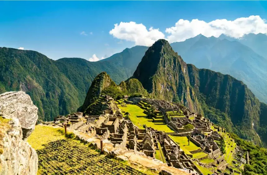 Vista panorâmica das antigas ruínas de pedra de Machu Picchu, com seus terraços agrícolas verdes espalhados pelo cume. Ao fundo, a icônica e íngreme montanha Huayna Picchu ergue-se majestosamente sob um céu azul com nuvens brancas, cercada por outros picos montanhosos da Cordilheira dos Andes.