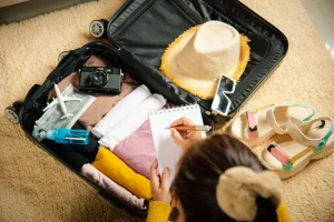 Vista de cima de uma pessoa escrevendo em um caderno de anotações enquanto prepara sua mala de viagem, que está aberta sobre um tapete felpudo bege. A mala preta contém roupas bem dobradas, uma câmera fotográfica estilo vintage, um chapéu de palha, óculos de sol brancos, uma máscara facial, um pequeno frasco azul e uma miniatura de avião. Ao lado da mala, no tapete, há um par de sandálias plataforma coloridas.