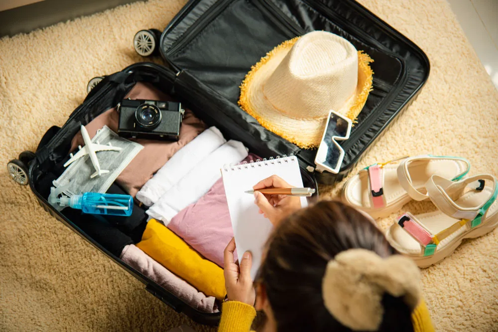 Vista de cima de uma pessoa escrevendo em um caderno de anotações enquanto prepara sua mala de viagem, que está aberta sobre um tapete felpudo bege. A mala preta contém roupas bem dobradas, uma câmera fotográfica estilo vintage, um chapéu de palha, óculos de sol brancos, uma máscara facial, um pequeno frasco azul e uma miniatura de avião. Ao lado da mala, no tapete, há um par de sandálias plataforma coloridas.