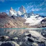 Uma vista deslumbrante de picos de montanhas rochosas e cobertas de neve (Monte Fitz Roy) refletidos perfeitamente nas águas cristalinas de um lago glacial. Em primeiro plano, pedras cinzentas repousam no fundo raso do lago sob um céu azul com nuvens.