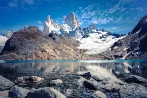 Uma vista deslumbrante de picos de montanhas rochosas e cobertas de neve (Monte Fitz Roy) refletidos perfeitamente nas águas cristalinas de um lago glacial. Em primeiro plano, pedras cinzentas repousam no fundo raso do lago sob um céu azul com nuvens.