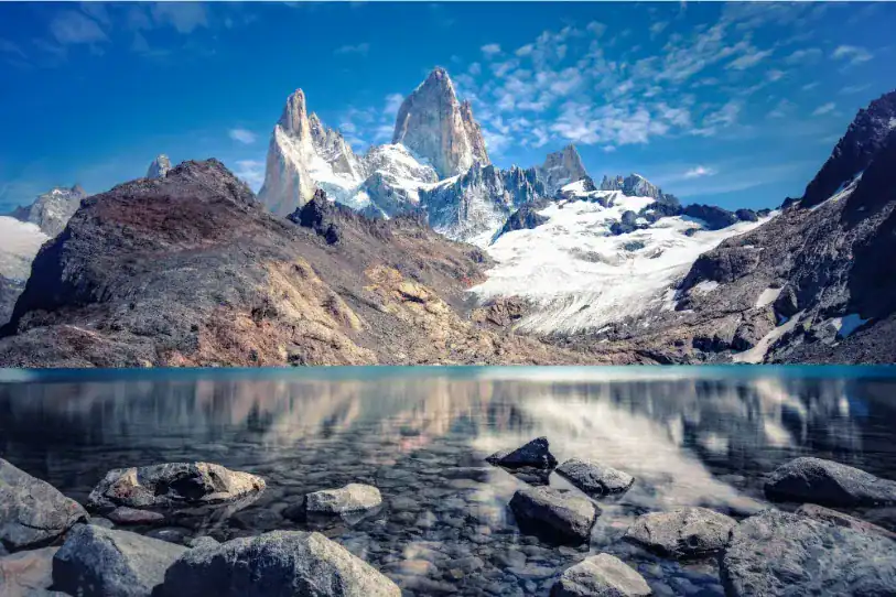 Uma vista deslumbrante de picos de montanhas rochosas e cobertas de neve (Monte Fitz Roy) refletidos perfeitamente nas águas cristalinas de um lago glacial. Em primeiro plano, pedras cinzentas repousam no fundo raso do lago sob um céu azul com nuvens.