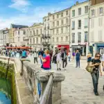 Uma orla de porto movimentada na Europa, com uma fileira de prédios históricos de pedra de múltiplos andares sob um céu azul com nuvens. O calçadão de paralelepípedos está repleto de pessoas caminhando, passeando e jantando em cafés ao ar livre. Uma mureta de pedra baixa corre ao longo da borda da água, que reflete a cena