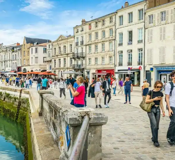 Uma orla de porto movimentada na Europa, com uma fileira de prédios históricos de pedra de múltiplos andares sob um céu azul com nuvens. O calçadão de paralelepípedos está repleto de pessoas caminhando, passeando e jantando em cafés ao ar livre. Uma mureta de pedra baixa corre ao longo da borda da água, que reflete a cena