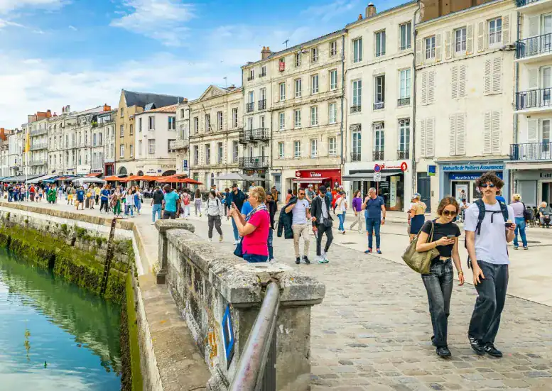 Uma orla de porto movimentada na Europa, com uma fileira de prédios históricos de pedra de múltiplos andares sob um céu azul com nuvens. O calçadão de paralelepípedos está repleto de pessoas caminhando, passeando e jantando em cafés ao ar livre. Uma mureta de pedra baixa corre ao longo da borda da água, que reflete a cena