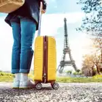 Pessoa puxando uma mala de viagem amarela em frente à Torre Eiffel, em Paris, durante um dia ensolarado.