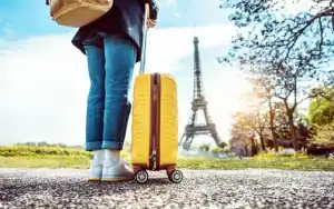 Pessoa puxando uma mala de viagem amarela em frente à Torre Eiffel, em Paris, durante um dia ensolarado.