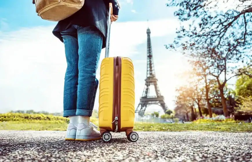 Pessoa puxando uma mala de viagem amarela em frente à Torre Eiffel, em Paris, durante um dia ensolarado.