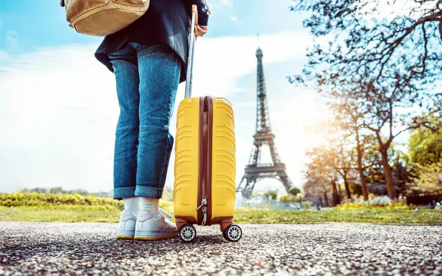 Pessoa puxando uma mala de viagem amarela em frente à Torre Eiffel, em Paris, durante um dia ensolarado.