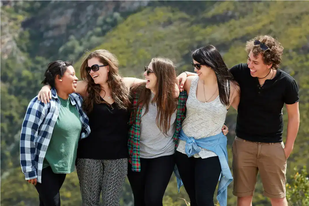Um grupo de cinco jovens amigos, sendo quatro mulheres e um homem, abraçados lado a lado e rindo de forma descontraída ao ar livre. Eles vestem roupas casuais e a maioria usa óculos de sol. Ao fundo, há uma encosta coberta por vegetação verde iluminada pelo sol, sugerindo um passeio na natureza ou trilha.