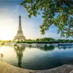 Uma vista panorâmica da Torre Eiffel em Paris durante o amanhecer ou entardecer. A torre se destaca no horizonte e seu reflexo é visível nas águas calmas do Rio Sena. A cena é emoldurada por grandes árvores verdes em ambos os lados, com calçadões de pedra ao longo das margens em primeiro plano. Vários barcos estão atracados ao longo do rio, sob um céu azul claro e iluminado pelo sol dourado à esquerda.