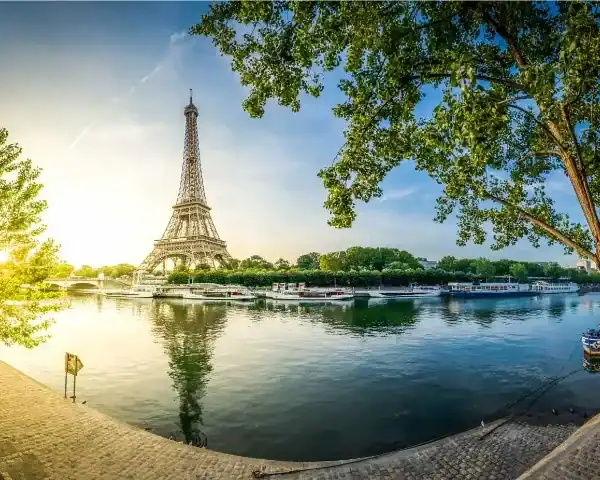 Uma vista panorâmica da Torre Eiffel em Paris durante o amanhecer ou entardecer. A torre se destaca no horizonte e seu reflexo é visível nas águas calmas do Rio Sena. A cena é emoldurada por grandes árvores verdes em ambos os lados, com calçadões de pedra ao longo das margens em primeiro plano. Vários barcos estão atracados ao longo do rio, sob um céu azul claro e iluminado pelo sol dourado à esquerda.