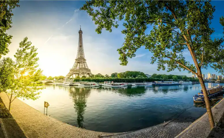 Uma vista panorâmica da Torre Eiffel em Paris durante o amanhecer ou entardecer. A torre se destaca no horizonte e seu reflexo é visível nas águas calmas do Rio Sena. A cena é emoldurada por grandes árvores verdes em ambos os lados, com calçadões de pedra ao longo das margens em primeiro plano. Vários barcos estão atracados ao longo do rio, sob um céu azul claro e iluminado pelo sol dourado à esquerda.