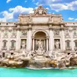 Uma vista frontal e majestosa da Fontana di Trevi, em Roma, sob um céu azul vibrante com nuvens brancas. A monumental fonte barroca de mármore claro destaca-se com suas esculturas detalhadas, centradas na imponente figura de Oceano sob um grande arco clássico. A água flui em cascata sobre formações rochosas esculpidas e deságua em uma ampla piscina de águas cristalinas em tons de azul-turquesa na base do monumento. Nas bordas laterais, é possível ver pedaços de edifícios urbanos clássicos em tons de amarelo e laranja.
