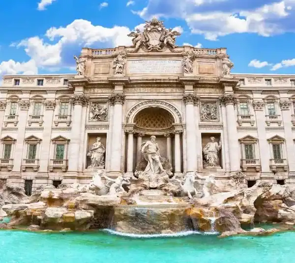 Uma vista frontal e majestosa da Fontana di Trevi, em Roma, sob um céu azul vibrante com nuvens brancas. A monumental fonte barroca de mármore claro destaca-se com suas esculturas detalhadas, centradas na imponente figura de Oceano sob um grande arco clássico. A água flui em cascata sobre formações rochosas esculpidas e deságua em uma ampla piscina de águas cristalinas em tons de azul-turquesa na base do monumento. Nas bordas laterais, é possível ver pedaços de edifícios urbanos clássicos em tons de amarelo e laranja.