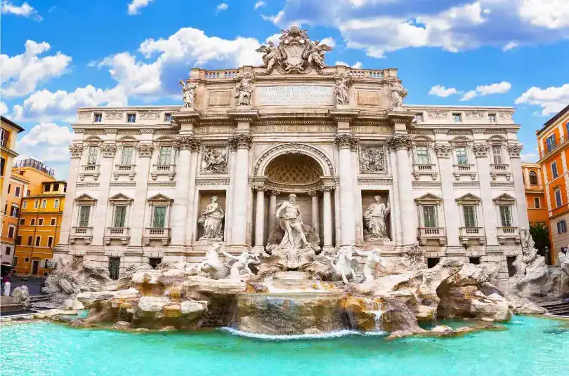 Uma vista frontal e majestosa da Fontana di Trevi, em Roma, sob um céu azul vibrante com nuvens brancas. A monumental fonte barroca de mármore claro destaca-se com suas esculturas detalhadas, centradas na imponente figura de Oceano sob um grande arco clássico. A água flui em cascata sobre formações rochosas esculpidas e deságua em uma ampla piscina de águas cristalinas em tons de azul-turquesa na base do monumento. Nas bordas laterais, é possível ver pedaços de edifícios urbanos clássicos em tons de amarelo e laranja.