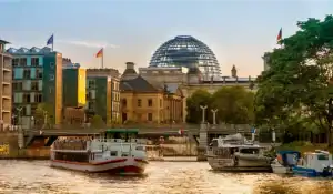 "Um barco navegando no rio Spree, em Berlim, durante o entardecer, com o edifício do Reichstag e sua icônica cúpula de vidro ao fundo."