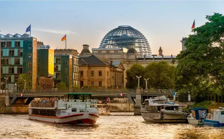 "Um barco navegando no rio Spree, em Berlim, durante o entardecer, com o edifício do Reichstag e sua icônica cúpula de vidro ao fundo."