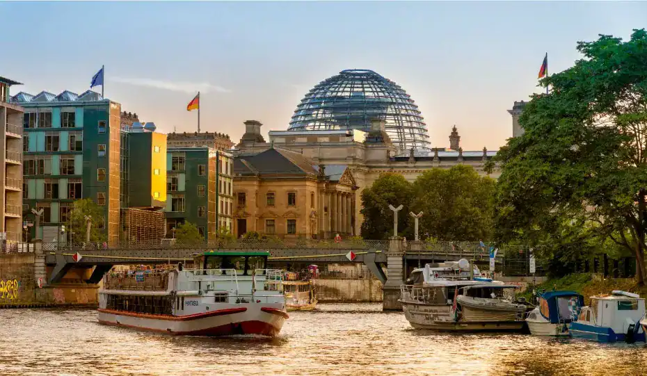 "Um barco navegando no rio Spree, em Berlim, durante o entardecer, com o edifício do Reichstag e sua icônica cúpula de vidro ao fundo."