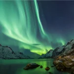 Vibrante aurora boreal verde sobre montanhas nevadas e um fiorde, com reflexos na água e rochas no primeiro plano.