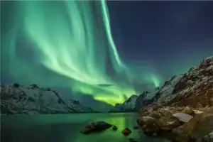 Vibrante aurora boreal verde sobre montanhas nevadas e um fiorde, com reflexos na água e rochas no primeiro plano.