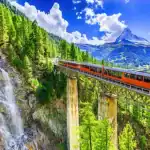 Trem laranja atravessando uma ponte sobre uma cachoeira, em meio às montanhas e florestas dos Alpes suíços, sob um céu azul com nuvens brancas.