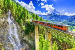 Trem laranja atravessando uma ponte sobre uma cachoeira, em meio às montanhas e florestas dos Alpes suíços, sob um céu azul com nuvens brancas.