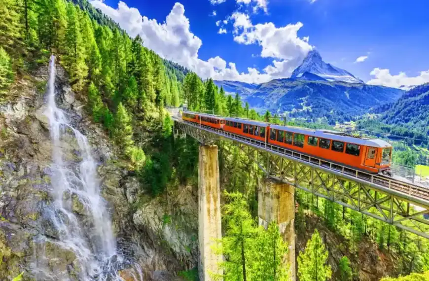 Trem laranja atravessando uma ponte sobre uma cachoeira, em meio às montanhas e florestas dos Alpes suíços, sob um céu azul com nuvens brancas.