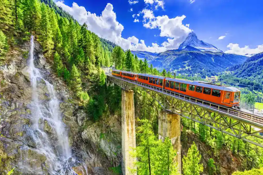Trem laranja atravessando uma ponte sobre uma cachoeira, em meio às montanhas e florestas dos Alpes suíços, sob um céu azul com nuvens brancas.