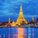 O Templo Wat Arun, na Tailândia, iluminado em tons dourados ao anoitecer, com seu reflexo brilhando nas águas do rio sob um céu azul escuro.