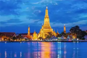 O Templo Wat Arun, na Tailândia, iluminado em tons dourados ao anoitecer, com seu reflexo brilhando nas águas do rio sob um céu azul escuro.
