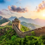 Fotografia da Grande Muralha da China a serpentear por montanhas cobertas de vegetação verde sob a luz do entardecer ou amanhecer. O sol brilha no lado direito, iluminando o céu com tons dourados e alaranjados entre as nuvens. Em destaque no centro, encontra-se uma torre de vigia de pedra, com a estrutura da muralha a estender-se continuamente pelos cumes das colinas ao fundo.