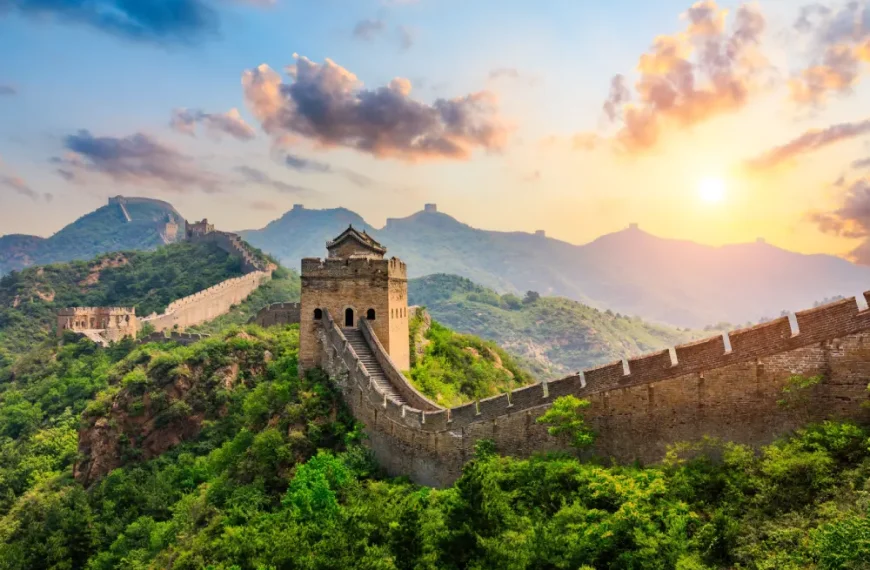 Fotografia da Grande Muralha da China a serpentear por montanhas cobertas de vegetação verde sob a luz do entardecer ou amanhecer. O sol brilha no lado direito, iluminando o céu com tons dourados e alaranjados entre as nuvens. Em destaque no centro, encontra-se uma torre de vigia de pedra, com a estrutura da muralha a estender-se continuamente pelos cumes das colinas ao fundo.