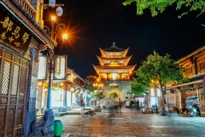 Rua histórica na China à noite, com arquitetura tradicional chinesa, lojas iluminadas por lanternas e um imponente pagode dourado ao fundo, cercado por árvores e construções típicas.