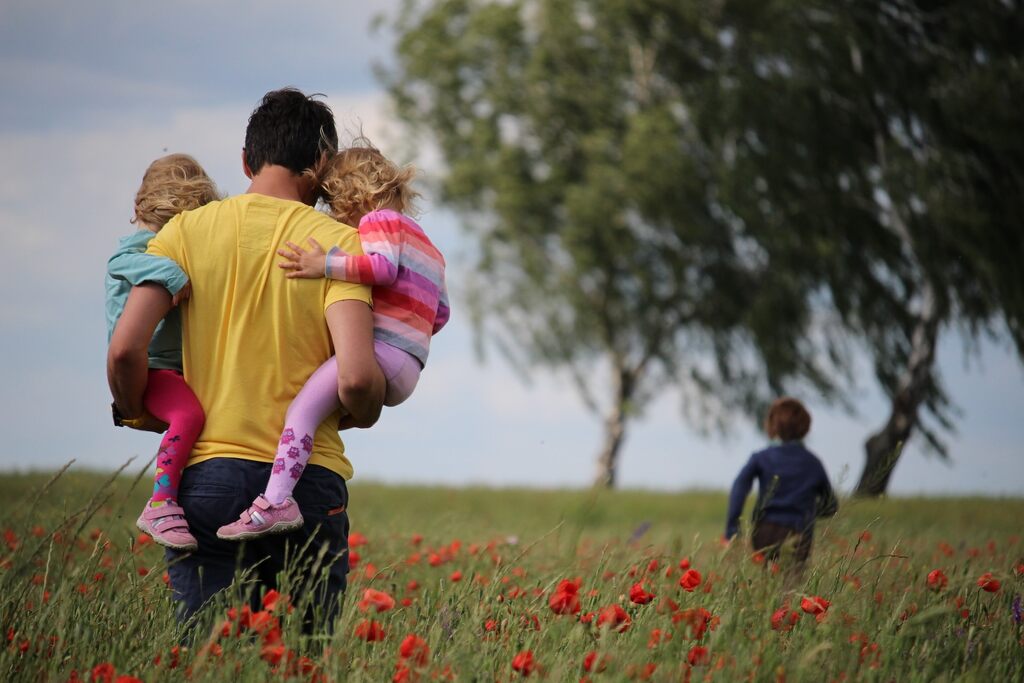 Viagem em família, pai segurando duas filhas e filho a frente brincando em um campo