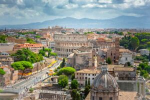 Vista panorâmica do centro de Roma, com o Coliseu ao fundo e ruas movimentadas com árvores verdes e edifícios históricos sob céu parcialmente nublado