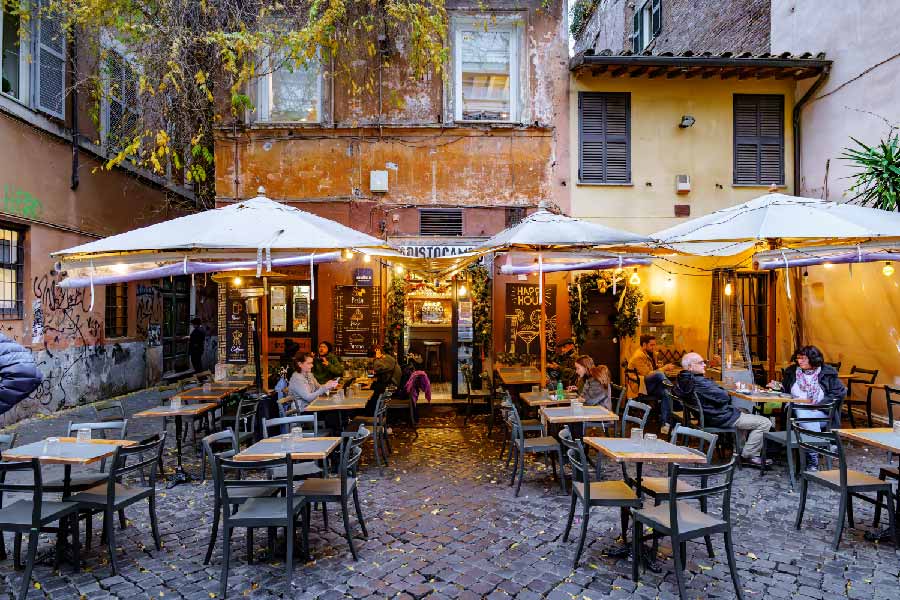 Cafeteria ao ar livre em uma rua de Roma, com mesas e cadeiras vazias, iluminação aconchegante e calçada de pedras, paredes com grafite e luminárias penduradas.